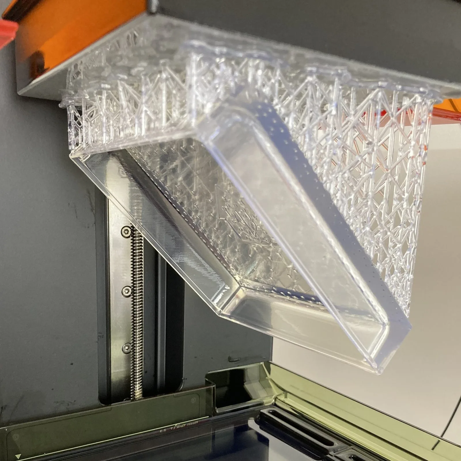 A clear prototype cover on the build plate of a resin 3D printer, still on its supports